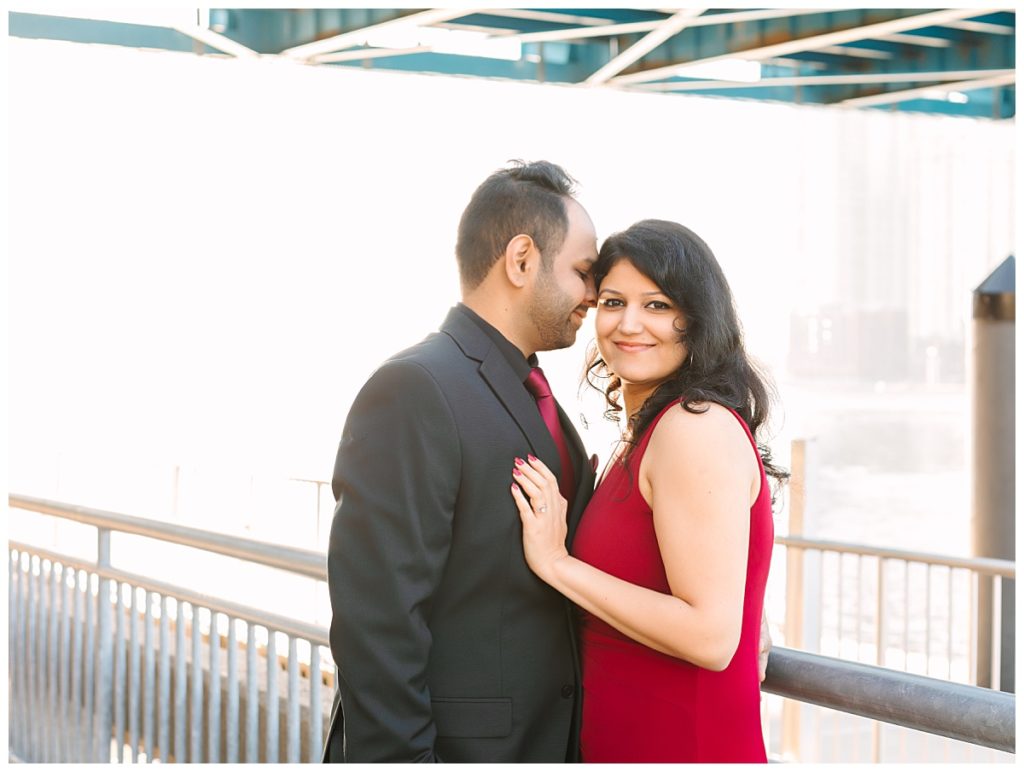 A dark haired couple embrace in a red dress and black suit for an outdoor engagement session in Jacksonville, Florida next to a bridge.