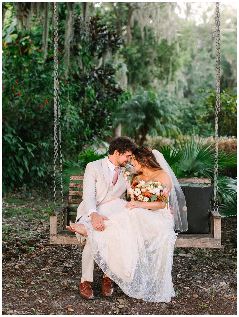 A dark haired groom with curls in a light tan tux embraces his barefoot brunette bride in a lace wedding gown on a porch swing at the Switzerland, FL wedding venue, La Venture Grove.