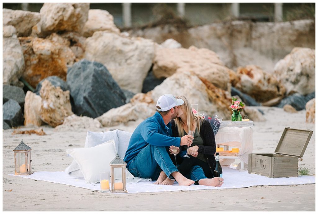 A newly engaged couple kiss on a picnic blanket on the beach in St. Augustine, FL