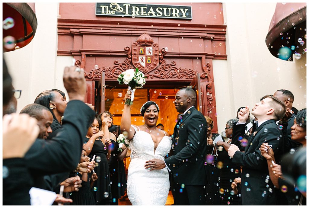 A bride in a white, lace gown with a plunging neckline holds her bouquet in the air next to her husband in a black tux surrounded by guests blowing bubbles outside The Treasury on Plaza.