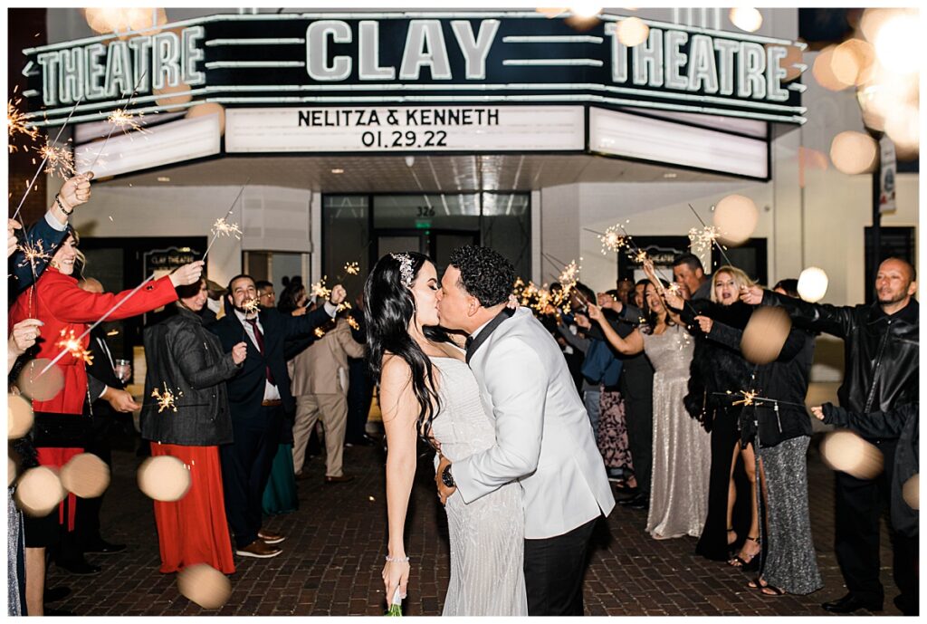 A newly married couple kiss under the marquee at The Clay Theatre surrounded by guests holding sparklers as they exit their wedding. 
