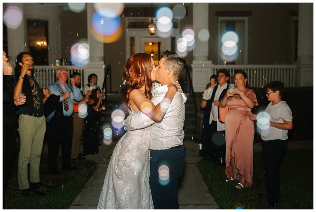 Florida newlyweds share a kiss as they exit their Jacksonville wedding reception surrounded by guests and whimsical bubbles.